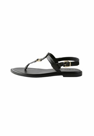 Sandalia de cuero negro con suela plana, diseño de tira en forma de T, acento de hardware en tono dorado y correa ajustable en el tobillo. Textura suave.