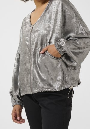 Kvinde iført skinnende sølvfarvet lynlåstrøje med elastiske manchetter og sorte bukser, hånd stukket ned i jakke lomme, udsnit af kroppen.