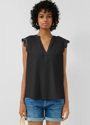 Vrouw met krullend haar, gekleed in een zwart mouwloos blouse met kanten randen op de schouders en blauwe opgerolde denim shorts, met een tas in haar hand.