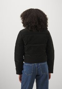 Sort pufferjakke med cropped design, quiltet tekstur og lange ærmer. Sammen med højtaljede blå jeans. Vist fra ryggen.
