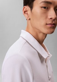 Mladý muž s krátkými tmavými vlasy, který má na sobě bílou polokošili s jemným šedým lemováním na límci, stojí mírně bokem.