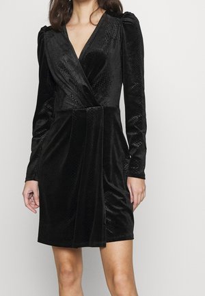 Femme portant une robe portefeuille en velours noir avec de longues manches bouffantes et un motif texturé rappelant un serpent, se tenant devant un fond clair uni.