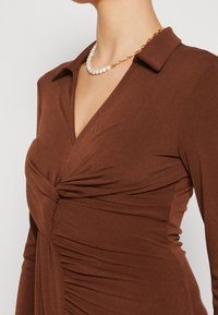 Femme portant une robe marron à manches longues avec un détail torsadé à l'avant et un collier combinant perles et maillons de chaîne dorés.