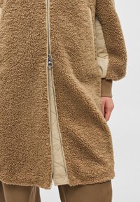 Brūns mākslīgās kažokādas mētelis ar rāvējslēdzēju priekšpusē, ar dūnu raksta bēšu oderi. Ietver kabatas un mīkstu, teksturētu ārpusi.