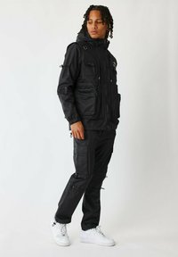 Zwarte waterdichte jas met meerdere voorzakken en verstelbare manchetten, gecombineerd met zwarte cargobroeken met ritsen en trekkoorden, gedragen met witte sneakers.