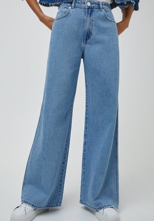 Jasnoniebieskie jeansy z wysokim stanem i szerokimi nogawkami, noszone z białymi sneakersami i krótką kurtką jeansową, ręce opierają się na biodrach.