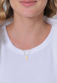 Gold cross pendant necklace on a thin chain, worn over a white ribbed top. The cross has a simple, polished design with a smooth texture.