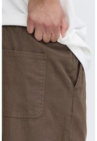 Pantaloni di cotone marroni con tasca posteriore e tasche sui lati, con una texture liscia e una vestibilità rilassata. La mano infila il tessuto sotto la camicia.
