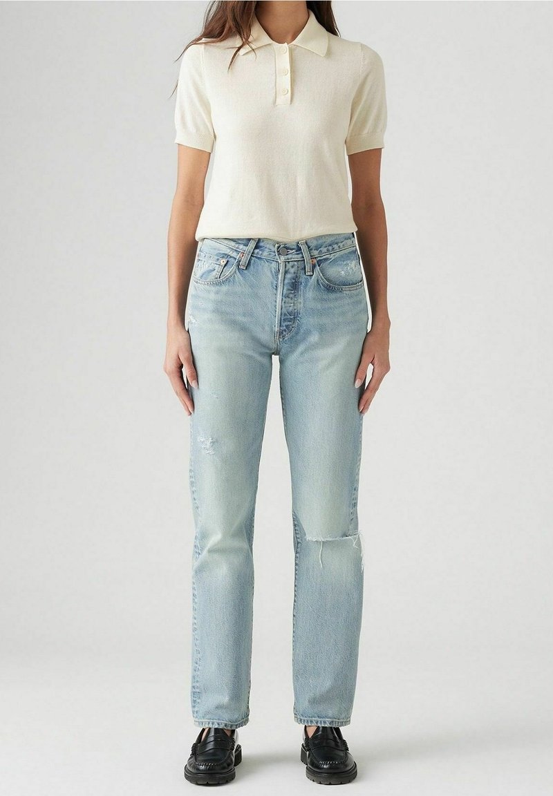Femme portant un polo crème à manches courtes, un jean taille haute déchiré bleu clair et des mocassins noirs, debout devant un fond uni.