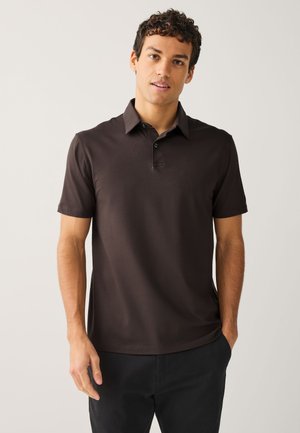 Mladý muž s kučeravými vlasmi, oblečený v tmavohnedej krátkorukávovej polokošeli a čiernych nohaviciach, stojaci pred jednofarebným svetlým pozadím.