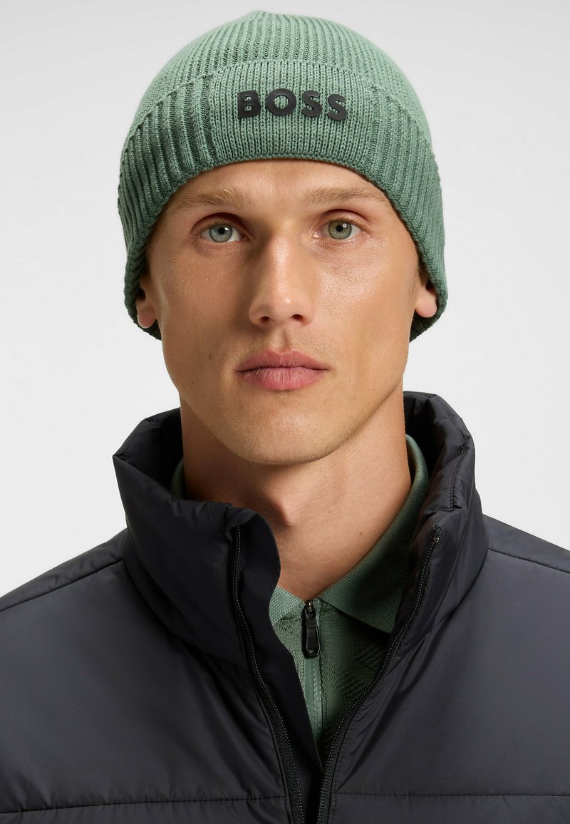 Green ribbed knit beanie with a black "BOSS" logo on the front. Worn with a black puffer jacket and a green polo shirt.