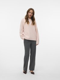 Frau steht mit lockerem, hellrosa Pullover, grauen geraden Hosen und braunen Spitzenschuhen vor einem weißen Hintergrund.