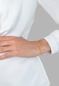 Zlata zapestnica z majhnimi okroglimi detajli, ki se ponaša z občutljivo zasnovo verige. Nosi se na zapestju in dopolnjuje belo oblačilo.