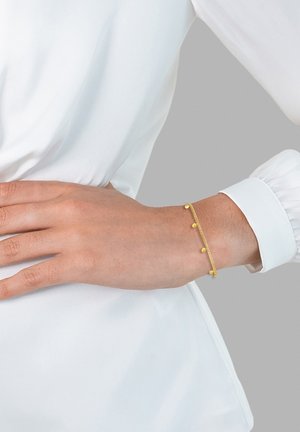 Pulsera de oro con pequeños acentos redondos, con un diseño de cadena delicado. Lleva puesta en una muñeca, complementando una prenda blanca.
