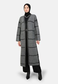 Lungo cardigan grigio con motivo a scacchi neri, dotato di maniche ampie, tasche laterali e taglio dritto. Indossato sopra un completo nero.