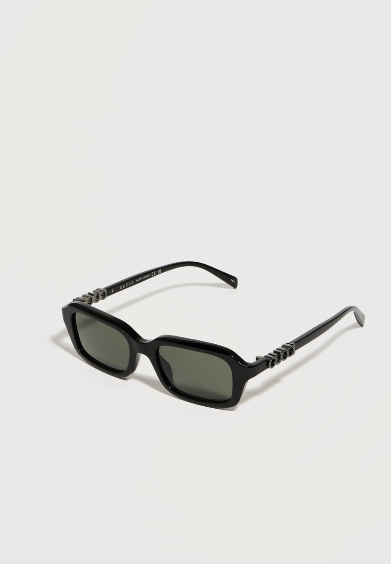 Lunettes de soleil rectangulaires noires avec des verres foncés et des logos Gucci argentés décoratifs sur les branches, présentées sur une surface blanche.