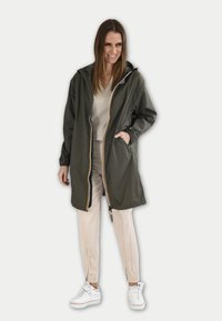 Olijfgroene lichtgewicht regenjas met een ritssluiting aan de voorkant, zijzakken en contrasterende gele afwerking. Draag over lichte broek en schoenen.