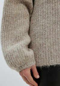 Beiger gestrickter Pullover aus weichem, strukturiertem Stoff. Mit lockeren Ärmeln und einem leicht ausgefransten Saum, der das gemütliche Design betont.