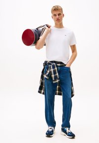 Biała koszulka, niebieskie dżinsy i niebieska koszula w kratę związana w talii. Trzyma okrągłą, czerwoną i granatową torbę podróżną. Czarnobiałe trampki.