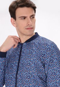 Geprinte jas met een geometrisch ontwerp in blauw, wit en rood. Het heeft een voorrits en een marineblauwe geribbelde kraag.