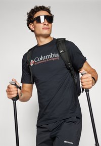 Černé tričko s krátkým rukávem s logem "Columbia Sportswear Company", ve spojení s tmavými kalhotami a slunečními brýlemi, držící trekové hole a nosící batoh.