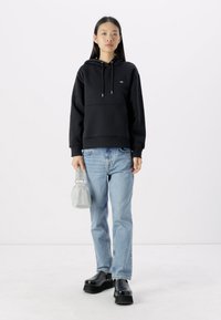Felpa nera con tasca frontale e cappuccio con cordini, abbinata a jeans azzurri chiari e scarpe con platform nere. Tiene una piccola borsa grigia.