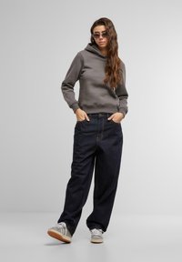 Un hoodie gris avec une poche kangourou à l'avant, associé à un jean bleu foncé ample. Des baskets grises avec des rayures blanches complètent la tenue.