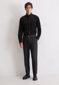 Junger Mann steht vor einer weißen Wand und trägt ein schwarzes Hemd mit Knöpfen, einen schwarzen Gürtel, dunkle graue Hosen und schwarze Schnürschuhe.