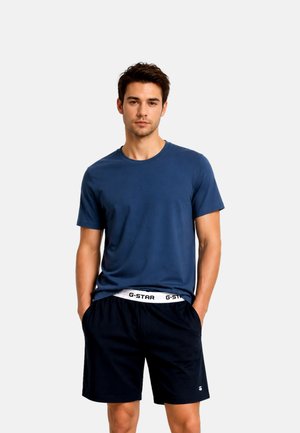 Jonge man staat met handen in zijn zakken, draagt een blauw T-shirt en zwarte korte broek met zichtbaar merklabel in de taille.
