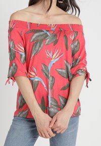 Top coral con escote descubierto y estampado floral que presenta hojas azules y verdes. Mangas cortas con detalle de lazo. Tejido ligero de mezcla de algodón.