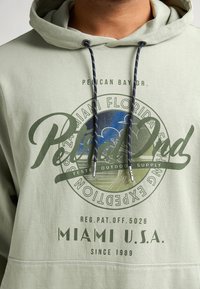 Sudadera con capucha de color verde claro con un gráfico circular que presenta "Pelican Bay Dr." y "Miami U.S.A." en verde oscuro. Cuerdas de ajuste en azul marino con puntas de plata.