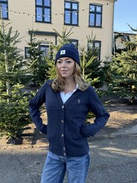 Mörkblå kabelstickad kofta med träknappar, med en liten logga på bröstet, kombinerad med en matchande stickad mössa och blå jeans.