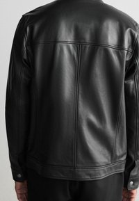 Schwarze Lederjacke mit glatter Textur, versehen mit einem runden Kragen, genähten Details und Druckknopfakzenten in der Nähe der Bündchen.