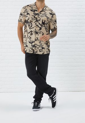 Beżowa koszula z krótkim rękawem z czarnymi wzorami smoka i fal, zestawiona z ciemnymi dżinsami i czarnymi trampkami z białymi paskami, stojąca na tle białej ceglanej ściany.