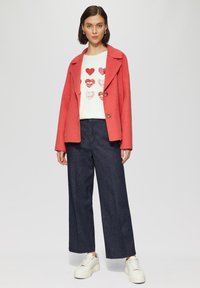 Koraal wollen mix jasje met een gekleed kraag, gecombineerd met een witte trui met rode hartjesprints en een donker denim wijde broek. Witte sneakers.