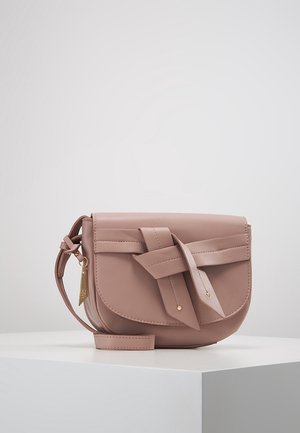 Bolso bandolera pequeño de cuero rosa empolvado con una solapa frontal ancha y detalle decorativo de correa anudada, colocado sobre una superficie blanca.