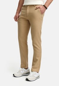 Pantalones beige de corte recto en una tela ligera, con parte delantera plana y bolsillos estándar, combinados con zapatillas deportivas gris y blanca.