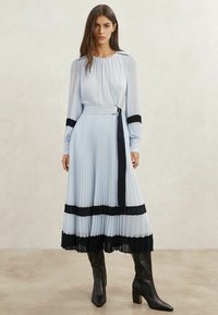 Rochie plisată bleu deschis cu accente negre și curea, având mâneci lungi și fustă fluidă. Asortată cu cizme negre până la genunchi.