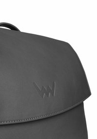 Šedý kožený batoh s klopovým zavíráním, vyraženým logem a detailem prošívání podél okrajů. Hladký povrch a strukturovaný design.