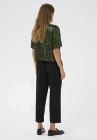 Vrouw met lang haar die een groen topje met korte mouwen en pailletjes, zwarte cropped broeken en zwarte loafers draagt, staande tegen een effen achtergrond.