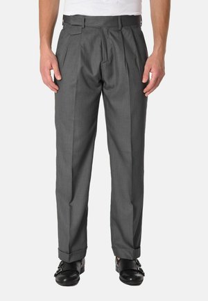 Uomo che indossa pantaloni grigi plissettati con orli risvoltati e scarpe nere double monk strap, in piedi davanti a uno sfondo semplice.