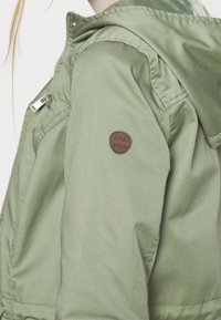 Persona che indossa una giacca leggera verde chiaro con un cappuccio, dotata di una toppa circolare marrone sulla manica con la scritta "ONLY SINCE 1995" e una zip frontale.