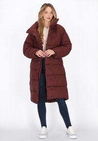 Giacca piumino bordeaux con colletto alto, bottoni a pressione e design trapuntato. Indossata sopra un maglione beige e jeans scuri, abbinata a sneakers bianche.