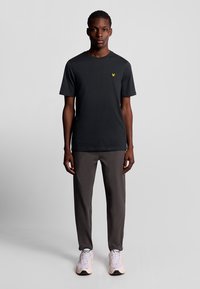 T-shirt nera in cotone con maniche corte e un piccolo logo giallo sul petto, abbinata a pantaloni grigi e sneaker chiare.
