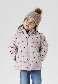 Giacca a vento imbottita di colore rosa con motivo a cuori neri, chiusura con zip e cappuccio. Indossata con un berretto a coste rosa con pompon in pelliccia.
