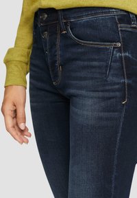 Tumed sinised denim teksad, millel on tuhmik tekstuur, esitaskud ja eristatav õmblus. Käsi puusal.