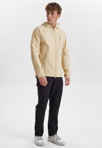 Vaaleankeltainen kevyt takki, jossa on huppu, vetoketjukfrontti ja sivutaskut, yhdistetty mustiin housuihin ja valkoisiin lenkkareihin. Suuri sileä pinta.