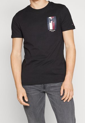 Homme portant un T-shirt noir à manches courtes avec un logo rectangulaire "H" multicolore et un texte sur la poitrine, associé à un jean gris.