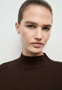 Suéter de punto marrón con cuello alto, que presenta una textura suave. Se puede ver el rostro de una modelo con un maquillaje minimalista y el cabello liso.