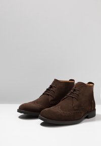 Paire de bottines brogues pour hommes en daim brun foncé avec semelles et lacets noirs sur une surface blanche et un fond gris.